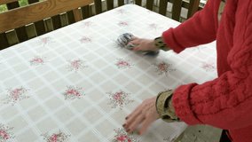 Senior elderly aged woman wipes and dusts dinner table on the patio outside the house. Country rural scene. 4K UHD video footage.