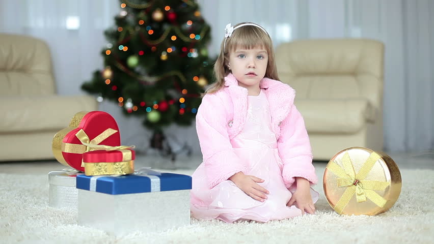 Child gets a gift from the box. She sits beside the Christmas tree. 