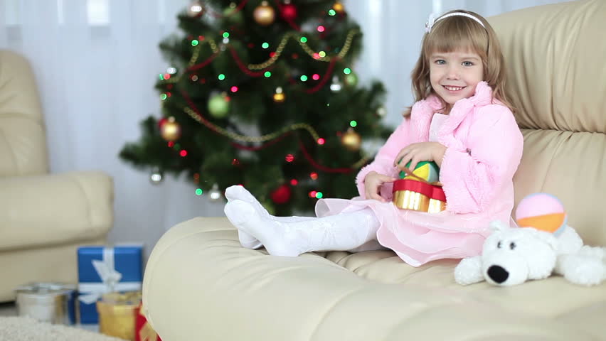 Happy little girl with toys sitting on the couch next to Christmas tree 