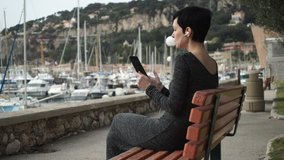  Female using app on  touchscreen modern device. Caucasian model happy to talking  with boyfriend at the vocation. She sitting on the bench at the seaside in mediterranean city nice provence 