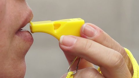Extreme close up shot of a woman blowing on a yellow whistle to make noise, give warning, get someone's attention or signal that something important is happening.