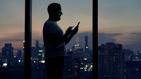 Young man using smartphone standing by window at night
