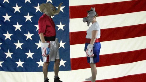 Man in elephant GOP mask and woman in donkey Democrat mask wearing boxing shorts sparring against against an American Flag .