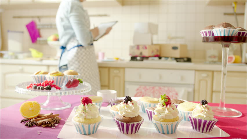 Woman counting boxes in the kitchen / Shot of tasty cup-cakes on the table in the fore ground out-of-focus, focused woman counting packs on the other table in the background 