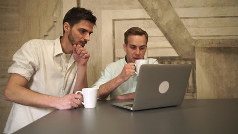 Friends drink coffee and tea consider the pros and cons of project. Mixed race man smiling. Caucasian man gesturing and point on the laptop monitor. 