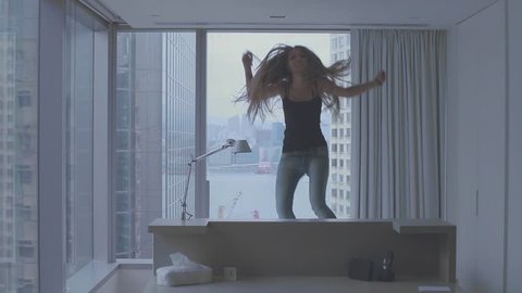 Young attractive teenage girl with long blonde hair having fun in the bed in a luxury apartment with breathtaking view over harbor in Hong Kong, China. Young woman jumping in the bed. Handheld shot.