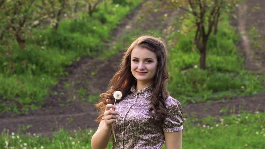 Young model blowing dandelion in the garden and smiling. Slowly