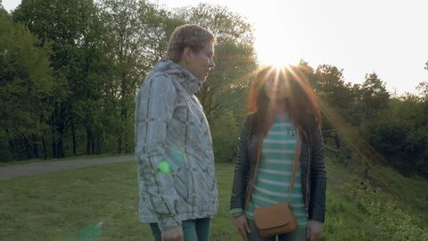 Two smiley merry and cheerful young women or girls friends walking in the green park outdoors in spring against the sunset. Togetherness and friendship positive concept. 4K UHD video footage.