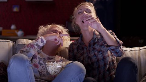 Two girls friends watching a movie on TV, eating popcorn and laugh