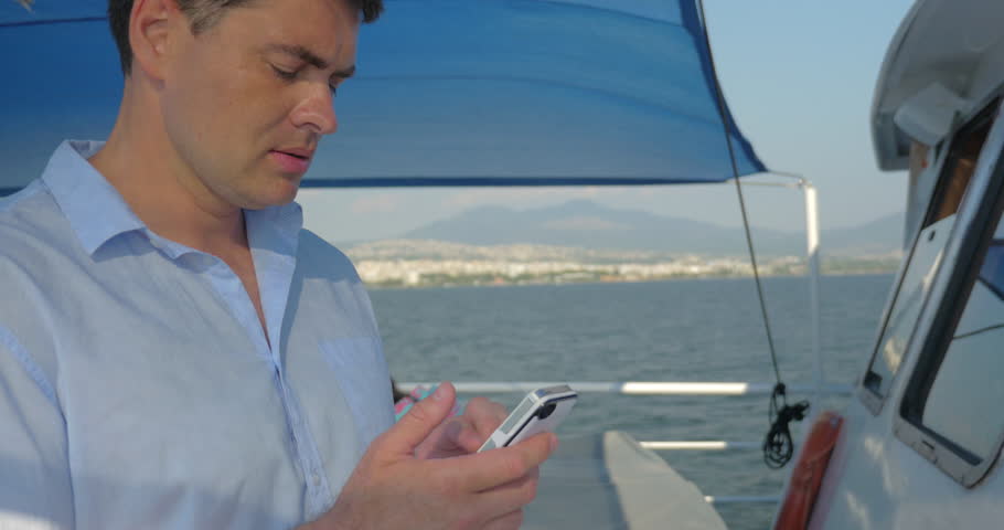 Young man typing on cellphone while traveling by ship. Sea and coastal city in background