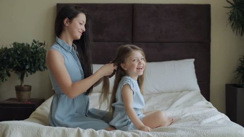A young mother sits with her daughter on the big bed, the mother braids the child's hair. Mom and daughter in blue dresses.