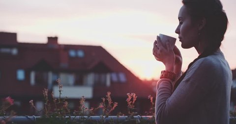 Woman Drinking Hot Coffee or Tea at the Balcony during Sunset. Slow Motion 120 fps, 4K DCi. Beautiful female enjoying cozy evening on terrace. Summertime relax at sunny patio flower garden. Lens Flare