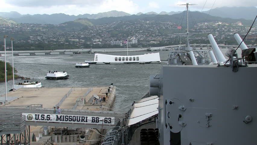 USS Missouri docked at Ford Island in Pearl Harbor, Honolulu, Oahu, Hawaii. Big deck guns aiming over USS Arizona memorial in a protective stance. Mountains in background. Entrance ramp and sign.