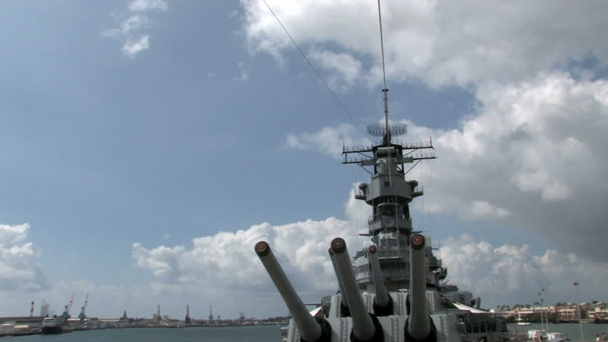 Missouri docked at Ford Island in Pearl Harbor, Honolulu, Oahu, Hawaii. Big deck guns aiming over camera view. Blue sky with clouds.