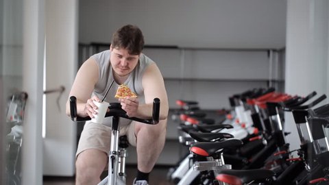 Fat guy turns pedals of exercise bike, in his hands he holds a drink and a slice of pizza