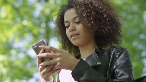 Portrait of relaxed young lady in a summer park reading a text message on her mobile phone. Beautiful young girl with dark curly hair using her cell phone, outdoor. Stock Video