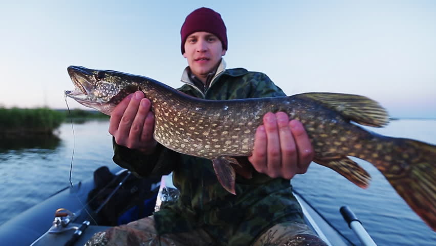 Happy young fisherman holding pike and sitting in the boat on the lake
