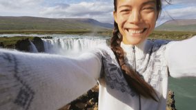 Tourist taking video selfie on travel by Godafoss waterfall on Iceland. Happy young woman tourists enjoying icelandic nature landscape visiting famous tourist destination attraction, Iceland