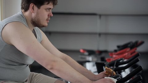 Tired man with an angry expression on his face eating pizza, engaging on an exercise bike