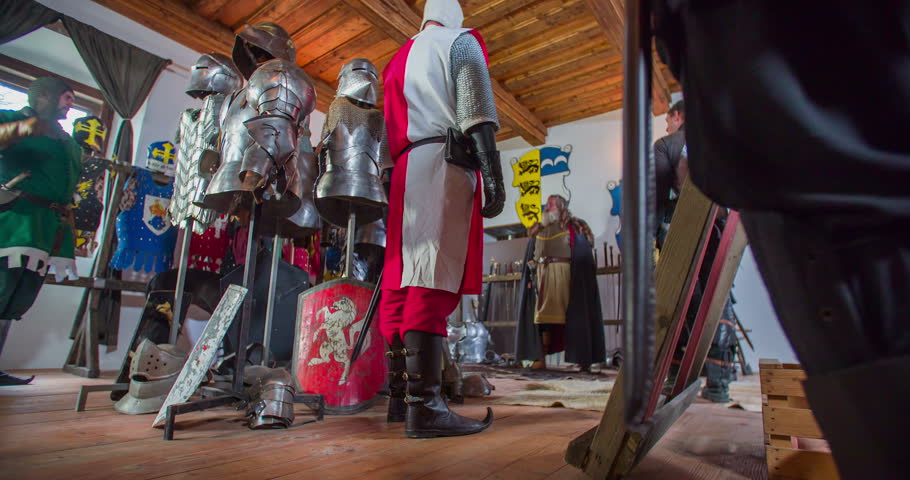 A commander finishes his speech and everyone lifts their hands up in agreement. Then he leaves the room, and the knights are getting dressed in armours and helmets. Wide-angle shot.