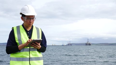 Asian engineer holding a tablet and standing in front of semi submersible oil rig in Invergordon Scotland
