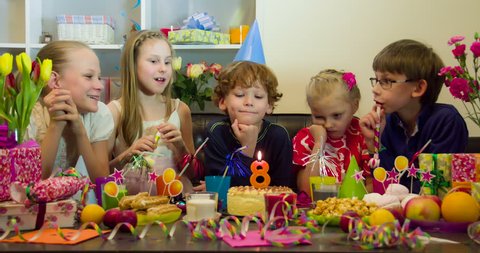 Joyful Birthday. Auburn curly-haired boy blows out the candle on a birthday cake. Together with friends he joyfully looking at serpentine falling