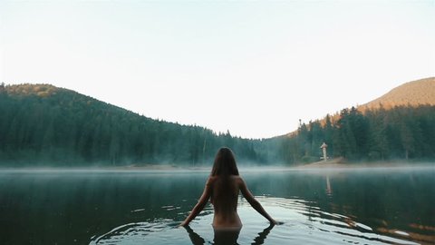 Beautiful naked fairy or mermaid with long dark hair in the mystical foggy mountain lake on back view. Fantasy magic world in mountains, videoclip de stoc