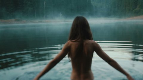Beautiful naked fairy or mermaid with long dark hair in the mystical foggy mountain lake on back view. Fantasy magic world in mountains