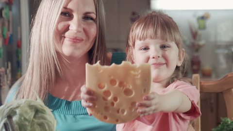 Beautiful mother and cute daughter at home in kitchen, biting tasting cheese and looking at camera and smiling. They are very happy and cheerful. 