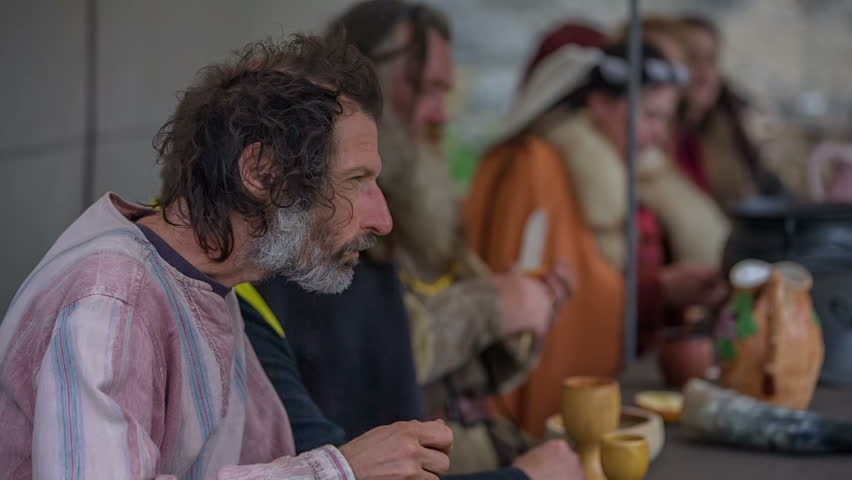 A royal family is eating dinner in a tent. On one side, there are men and on the other there are women and they are watching a fencing tournament. Close-up shot.