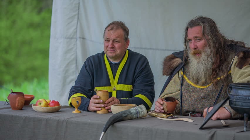 A king and a member of a royal family are talking to each other. They are also eating at a table and drinking some wine. Wide-angle shot.