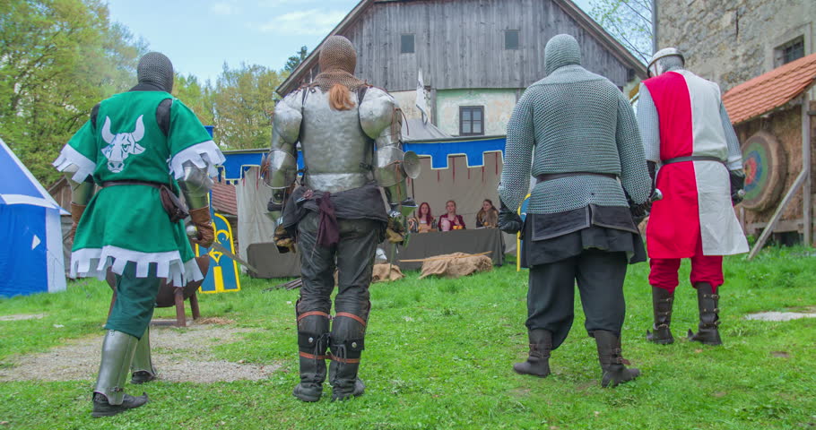 Four knights are walking up to their king who is sitting in a tent together with his family and they kneel down in front of them. The king lifts his cup up. Wide-angle shot.