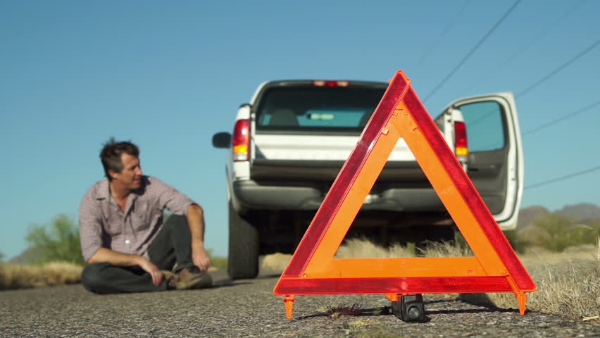 Male in the distance dramatically lays down on the road with his broken down truck with hazard lights on parked on the side of the road and an emergency marker reflective triangle in the foreground. Royalty-Free Stock Footage #16679422
