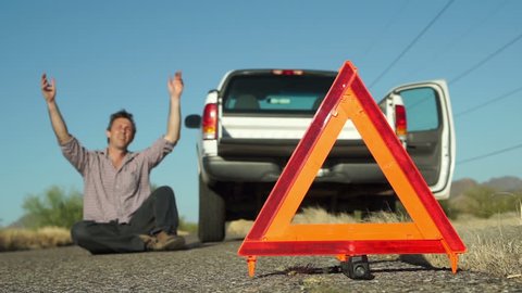 Male in the distance dramatically lays down on the road with his broken down truck with hazard lights on parked on the side of the road and an emergency marker reflective triangle in the foreground.