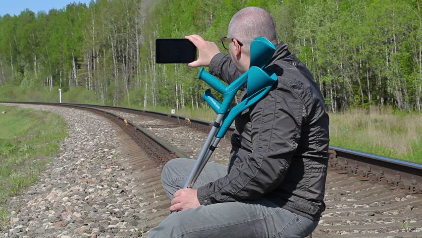 Disabled man with crutches sitting on railway and take pictures on smart phone
