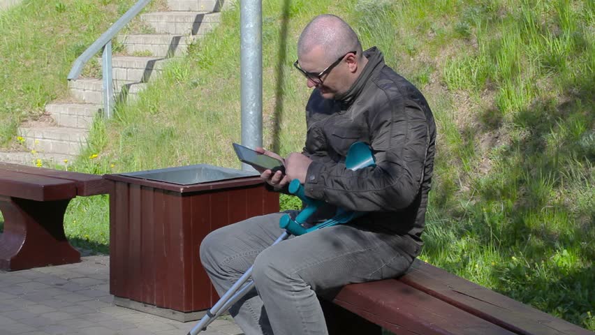 Disabled man with crutches sitting on bench and using tablet PC
