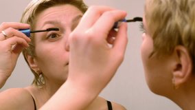 Pretty blond woman applying mascara make-up on her eyelashes in front of mirror and looks at herself. Beauty and makeup concept. 4K UHD video footage.