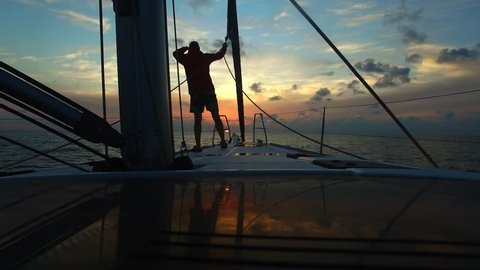 Young man standing with his back on the bow sailing yacht and looks forward in the course of the boat. Beautiful sunset sky in background. Look into the distance. Standing at deck of boat.