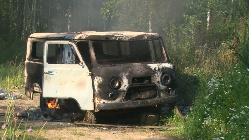 Old burning car emitting smoke in an empty summer sunny green field