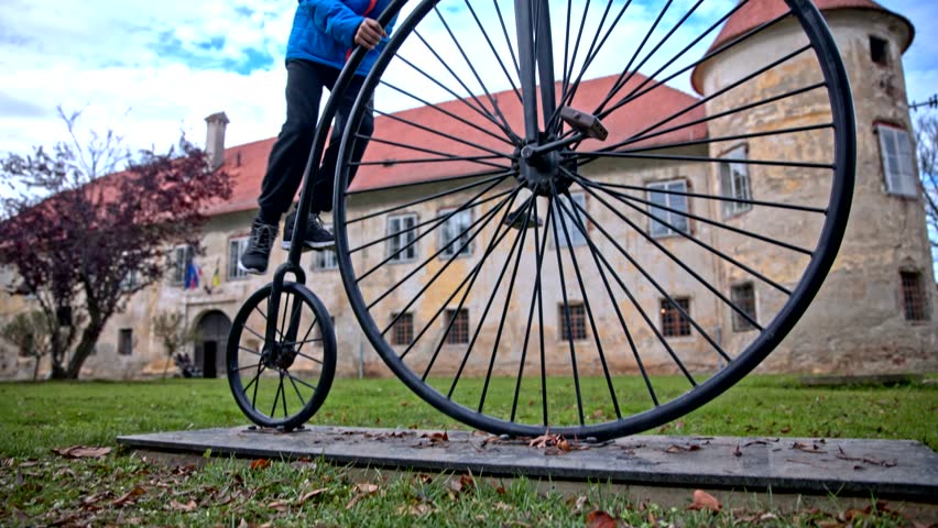 Boy climb to penny-farthing bicycle statue. Low angle slide of young person in blue jacket climbing to statue of very old bicycle with big front wheel. Old castle in background.