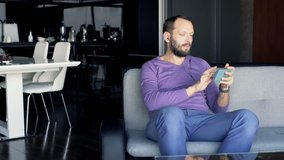 Young happy man watching movie on smartphone sitting on sofa at home 
