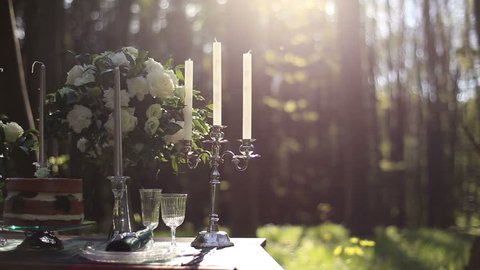 Close up of white vintage candelabrum on the wedding decorated table in the forest. Bouquet of white roses on background. Romantic dinner in the forest