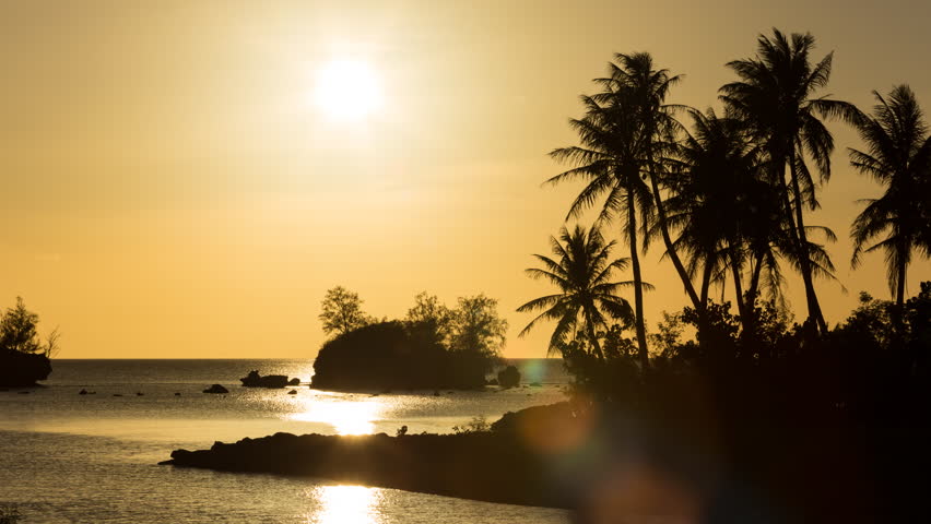 Palm trees and Islands off the coast of Guam are silhouetted by the setting sun through thinly veiled clouds that turn orange as the sun disappears. Agat, Guam