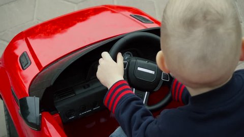 Baby Blonde Behind the Wheel of a Toy Car, a Boy Sitting in a Car Red Toy Car For Children, a Child Rides in the Park, a Beautiful Blonde, Fast Rides, 4k