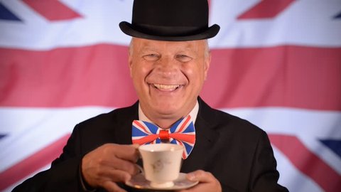 Laughing British businessman, takes a sip of tea from the cup, whilst the Union Jack flag blows in the background.