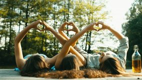 Three young Caucasian teen girls lying on a wooden pier, making heart shapes with their hands. Summer holidays, friendship concept. 60 FPS slow motion shot. Blackmagic URSA Mini RAW graded footage