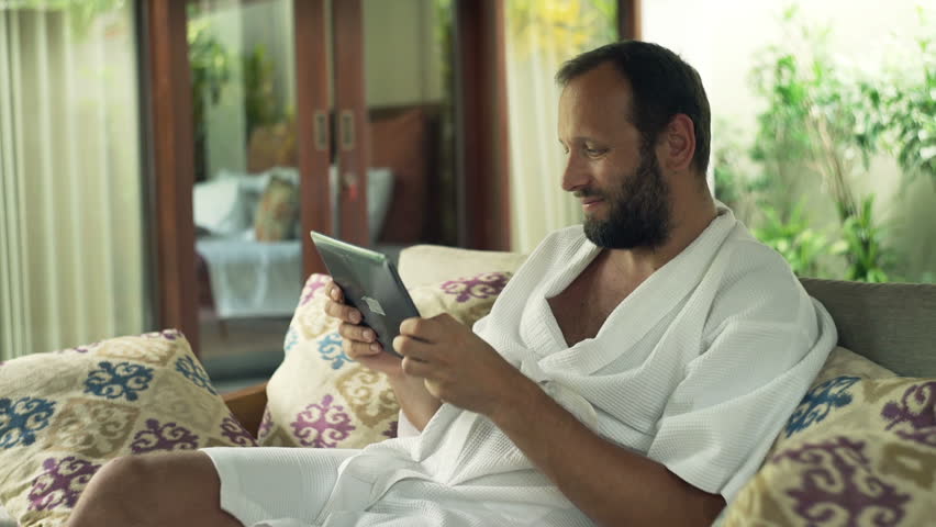 Young man in bathrobe playing game on tablet computer while sitting on sofa in outdoor villa

