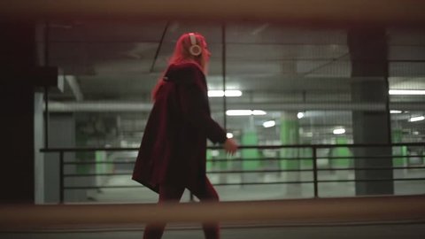 Teenage girl with long blonde hair walking fast along the street,listening to the music,wearing headphones at night under red lights, wearing black coat, jeans and high heel boots. 
