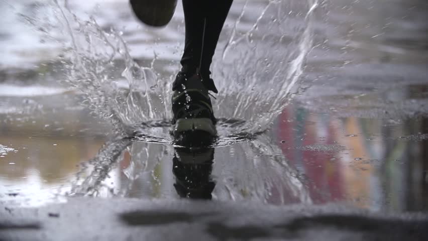 Close up slow motion shot of legs of a runner in sneakers. Male sports man jogging outdoors in a park, stepping into muddy puddle. Single runner running in rain, making splash.