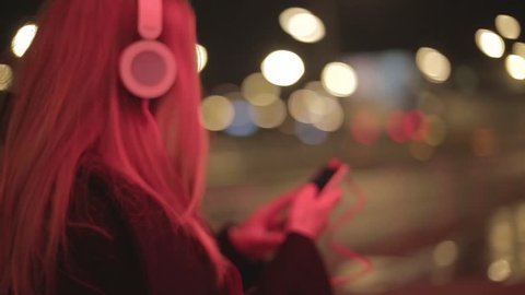 Teenage girl with long blonde hair walking along the street,listening to the music,dancing, smiling, wearing headphones at night under red lights, wearing black coat. Tracking shot.